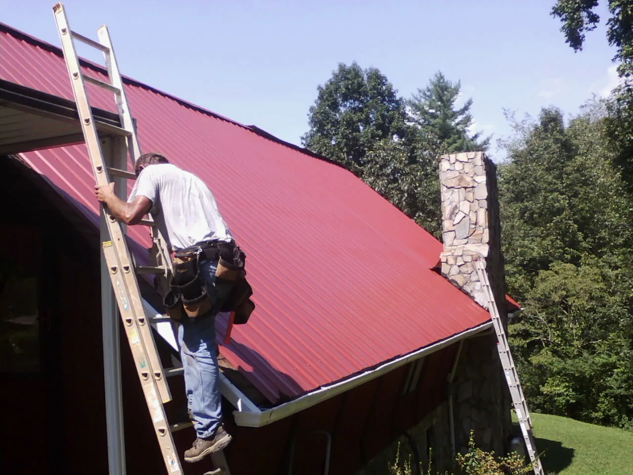 Roofer on a ladder beside a red metal roof and stone chimney — Soffit & Fascia Repair by Roofing Brothers in Kanaranzi